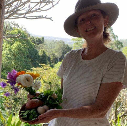 Jen Parke stands holding a bowl of fresh produce and a bunch of flowers smiling